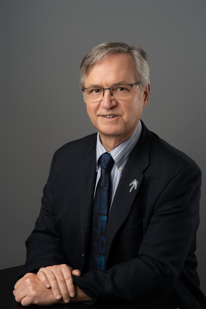 Professional headshot portrait of BC Treaty Comissioner George Abbott sitting down with his right hand on top of his left hand on a table