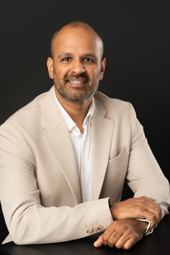 Yonel Prasad, Director of Strategic Partnership, sitting down for his corporate headshot and posed with right hand over left hand on table.