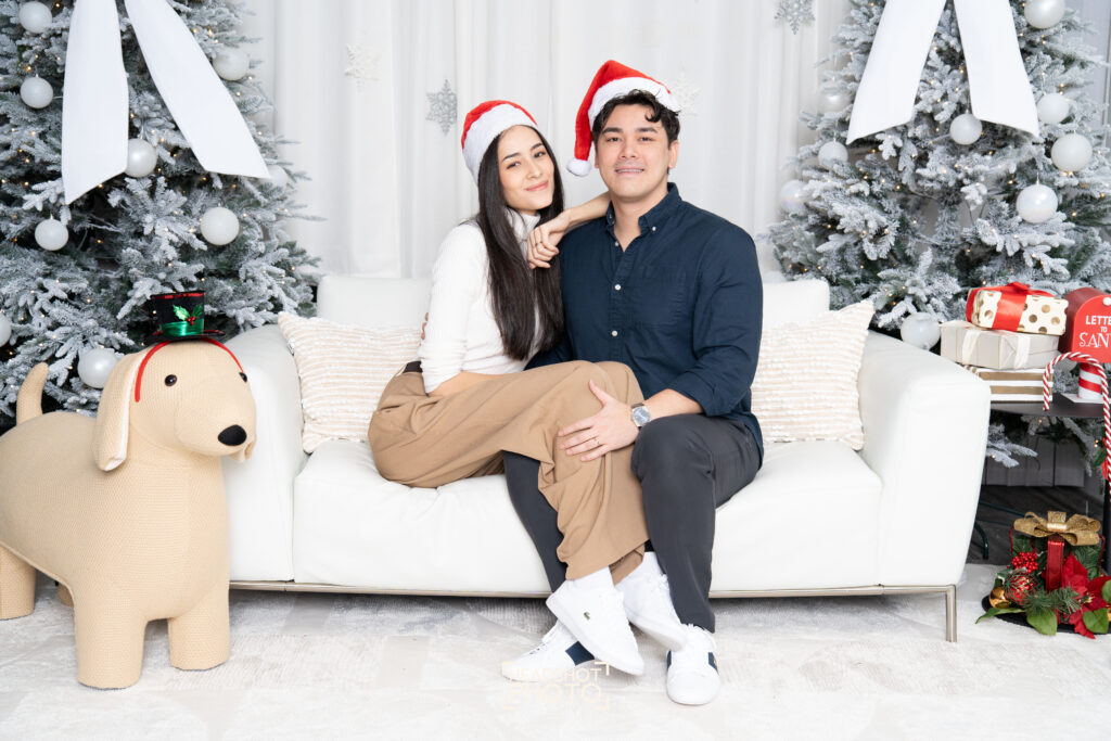 A couple wearing Santa hats posing on a white couch for a Christmas portrait session.